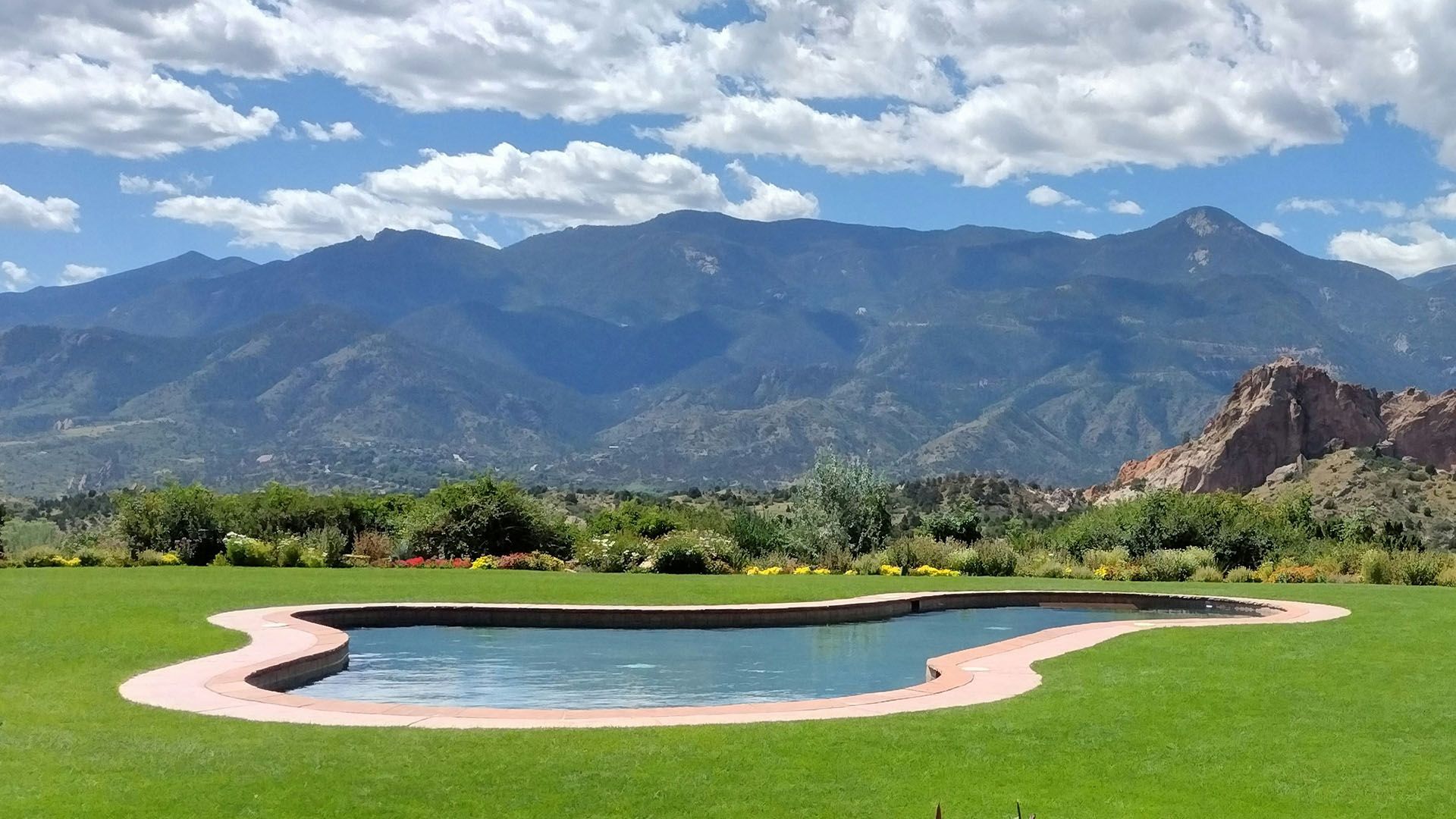 A scenic in-ground swimming pool is surrounded by a green lawn, with mountains in the background.
