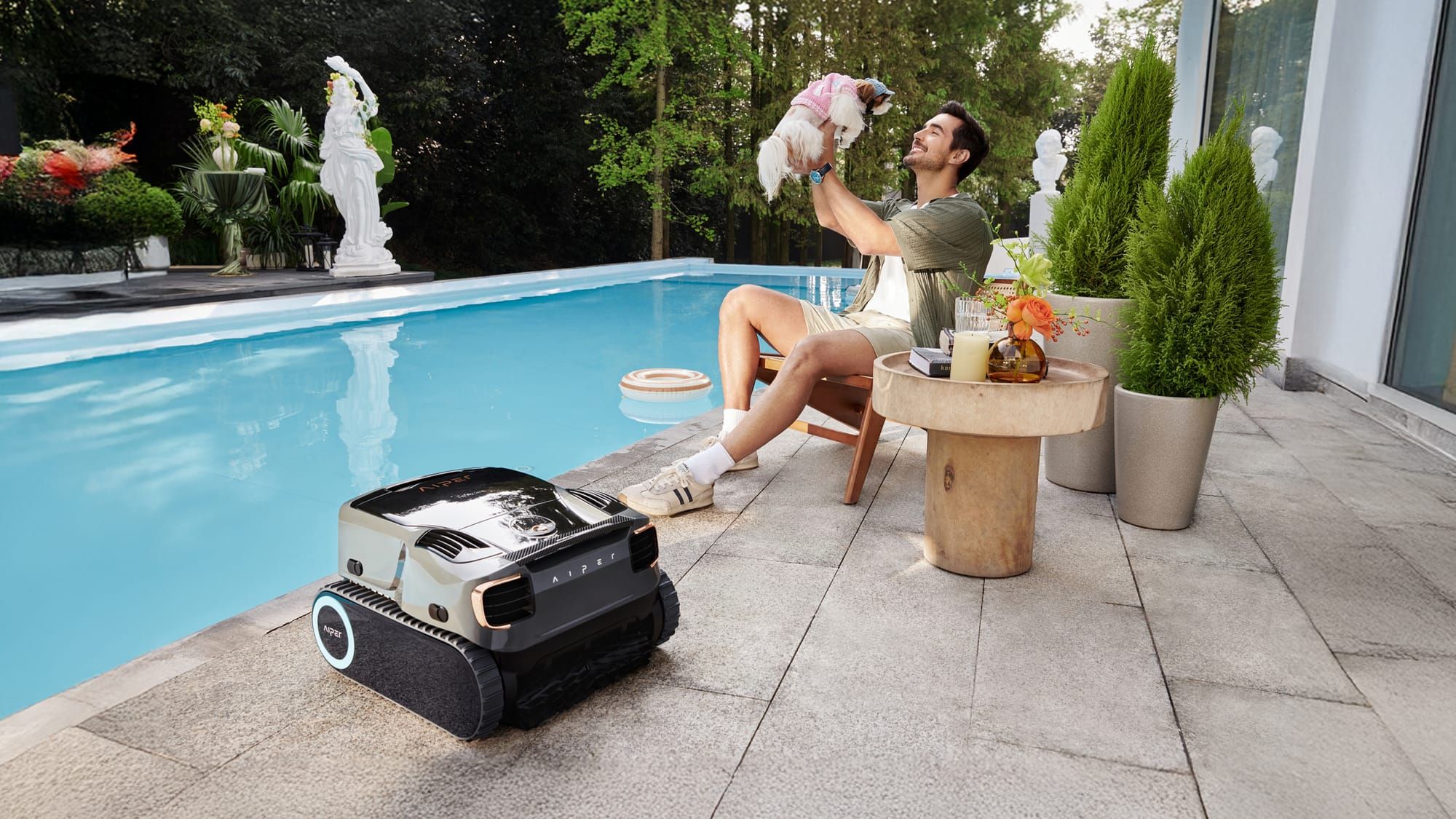 A man sits by a pool, happily holding a small dog, while a sleek robotic pool cleaner rests nearby on the patio.