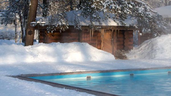 A steaming, heated outdoor pool in a snowy, winter landscape with a small cabin behind it.