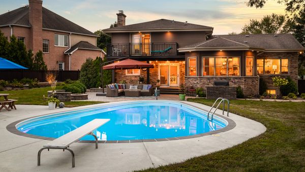 An illuminated inground pool with a diving board and patio furniture in a large backyard at dusk.