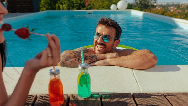 A man in sunglasses lounges in a pool, smiling at a woman with drinks on a new pool deck.