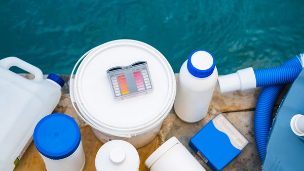 A variety of pool maintenance chemicals, a test kit, and a vacuum hose next to a swimming pool.