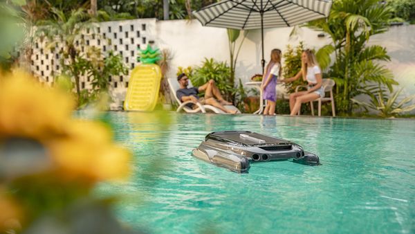 A pool automation robot skims the surface of a sunny swimming pool while a family relaxes nearby.