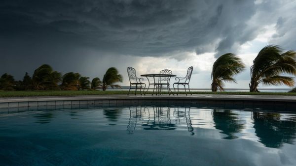 Dark storm clouds and strong wind over a backyard swimming pool with patio furniture, signaling an approaching hurricane.