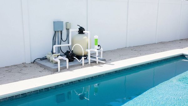 A pool sand filter is placed next to the swimming pool with a white wall behind it.