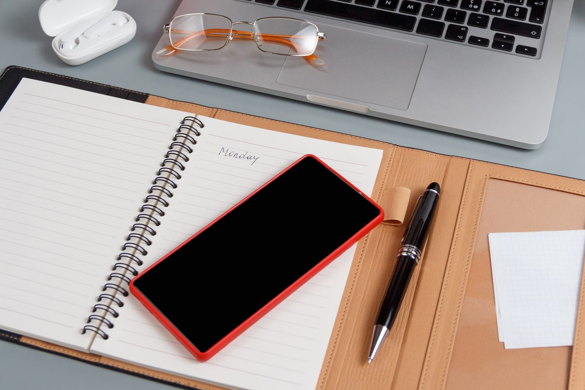Desk flatlay with a notebook opened written Monday on it, a mobile phone and a pen next to it, on top you see the corner of a laptop, earphones and a pair of glasses.