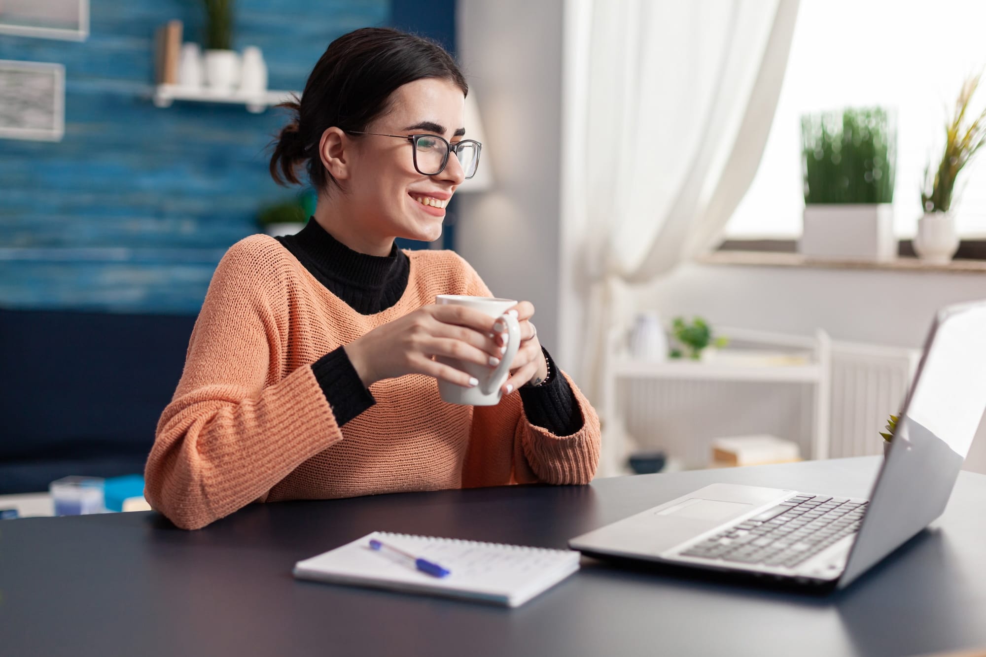 Female with brown hair, sitting on a desk, wearing large glasses and an orange sweater, she is holding a mug and smiling as she stares at her laptop screen.