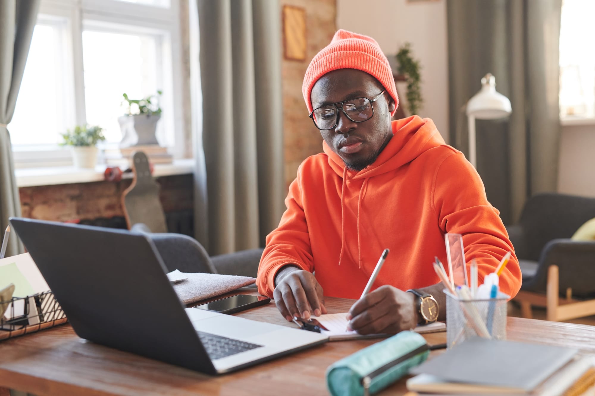Male wearing large frame glasses, orange knitted hat and hooded sweater, sitting on a desk, writing on a notepad with his laptop opened.